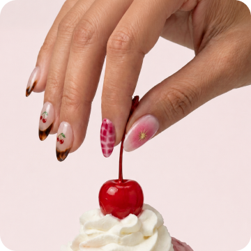 Close-up of a hand with Dashing Diva Ciao Ciao Magic Press Design, reaching for a cherry atop a whipped cream dessert against a pink background.