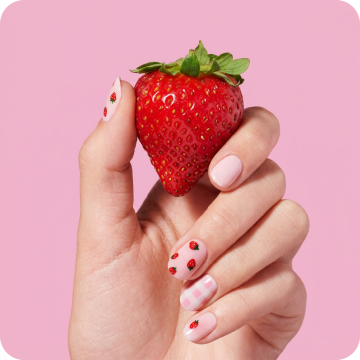 Close-up of a hand with Dashing Diva Central Park Picnic Glaze Design nails, holding a fresh strawberry against a pink background.