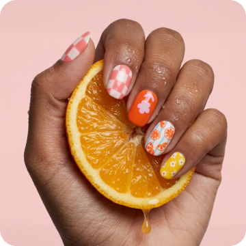 Close-up of a hand with Dashing Diva Buda-zest Glaze Design nails, holding a sliced orange against a pink background.