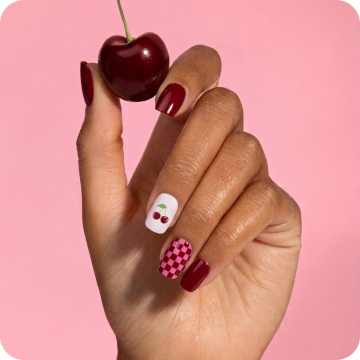 Close-up of a hand with Dashing Diva Cha Cha Cherry Gloss Palette nails, holding a fresh cherry against a pink background.
