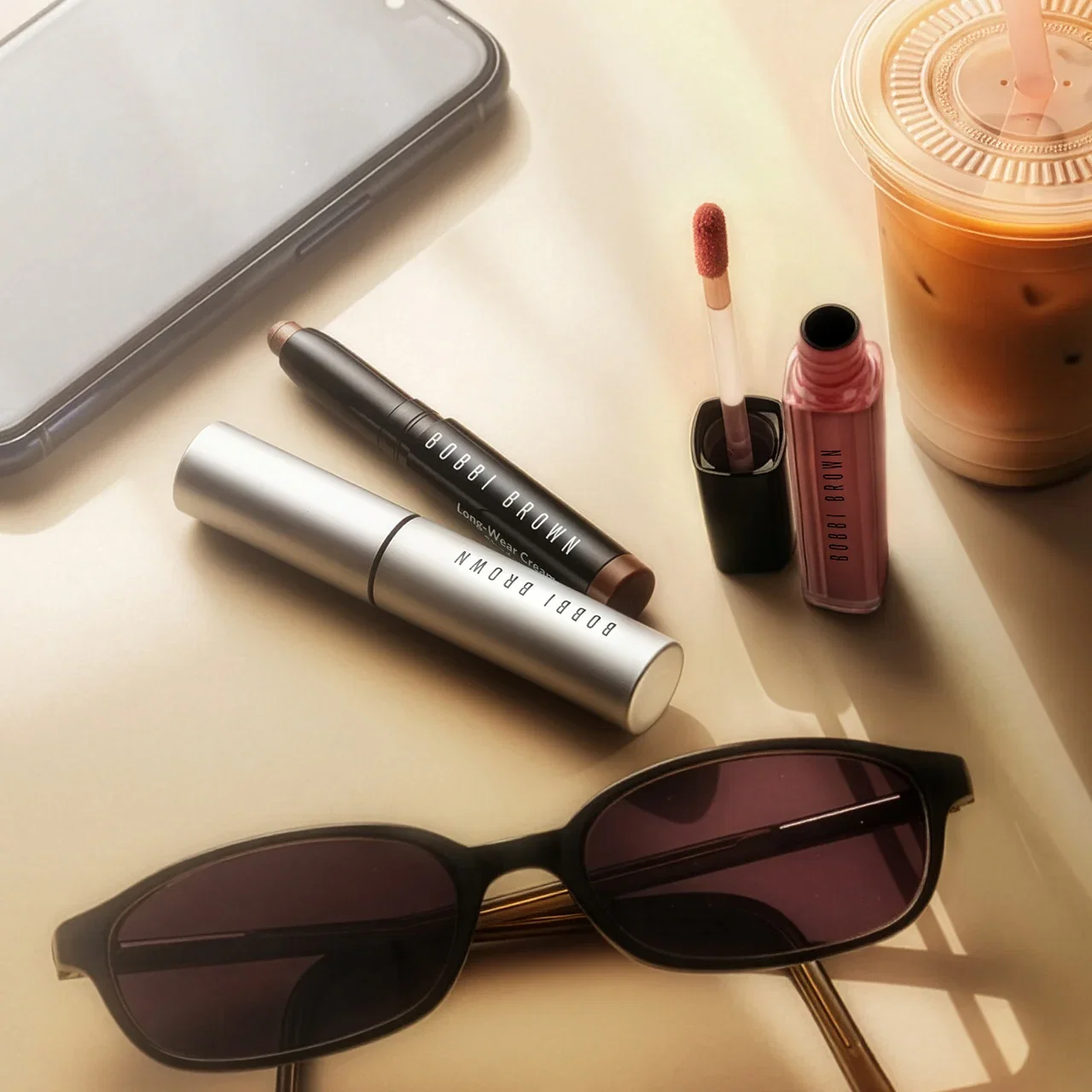 Flat lay of four Bobbi Brown mascara, eyeshadow sticks, and lip colour, with coffee, and sunglasses.