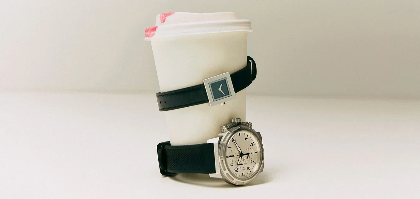 Vaso de café desechable con una mancha de lápiz labial, envuelto con dos relojes de pulsera sobre un fondo blanco liso.