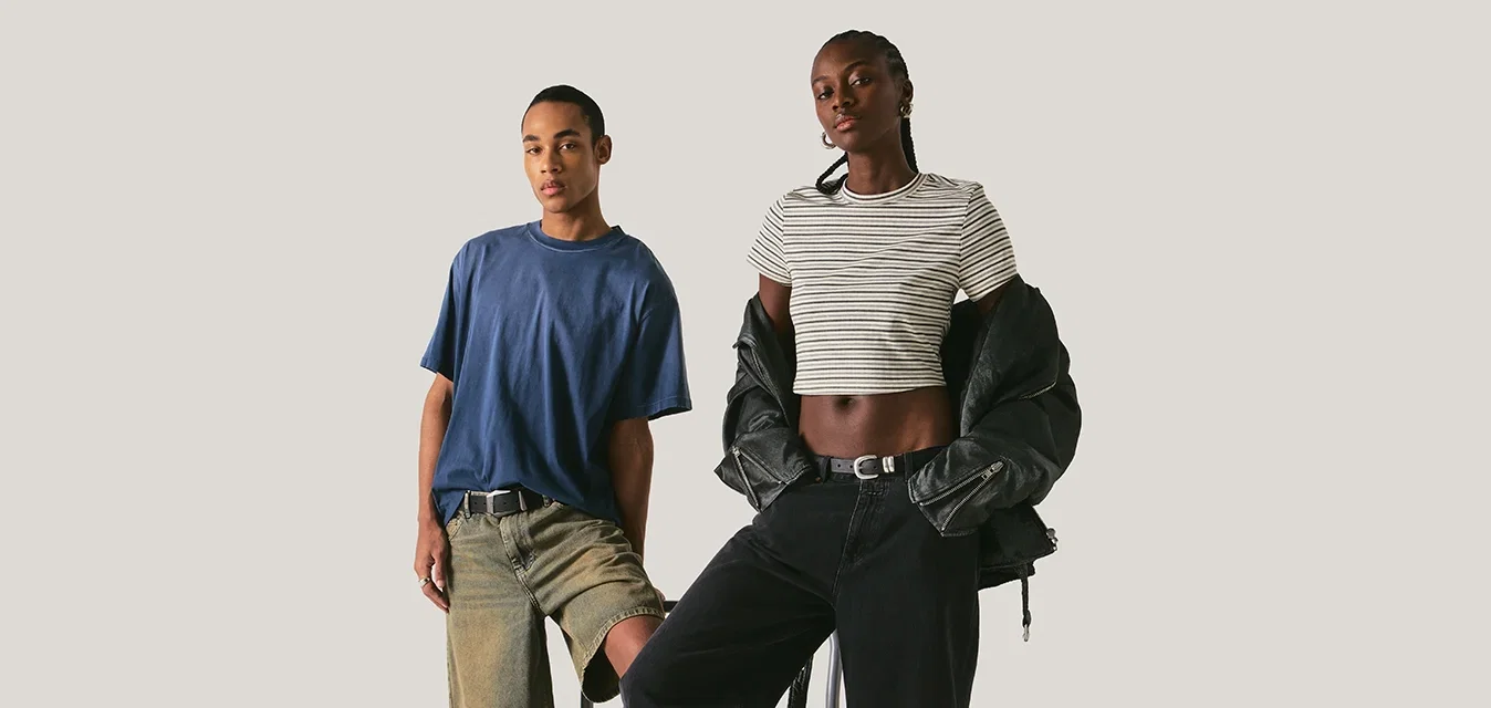 Dos personas posan con un carrito plano; una está de pie sobre él, vestida con pantalones cortos y botas, y la otra se apoya en el carrito, llevando un top corto de rayas y pantalones negros.