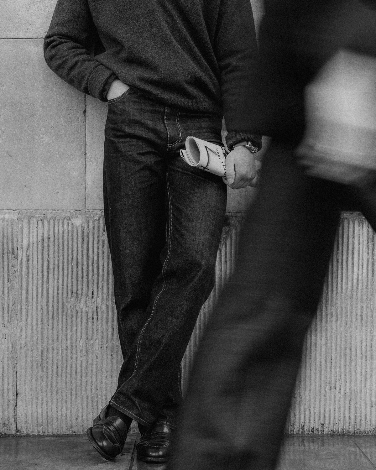 A black and white image of man leaning against wall wearing grey woold jumper, the Hack Selvedge jeans, and black leather shoes, holding a newspaper. A blurred person walks by.