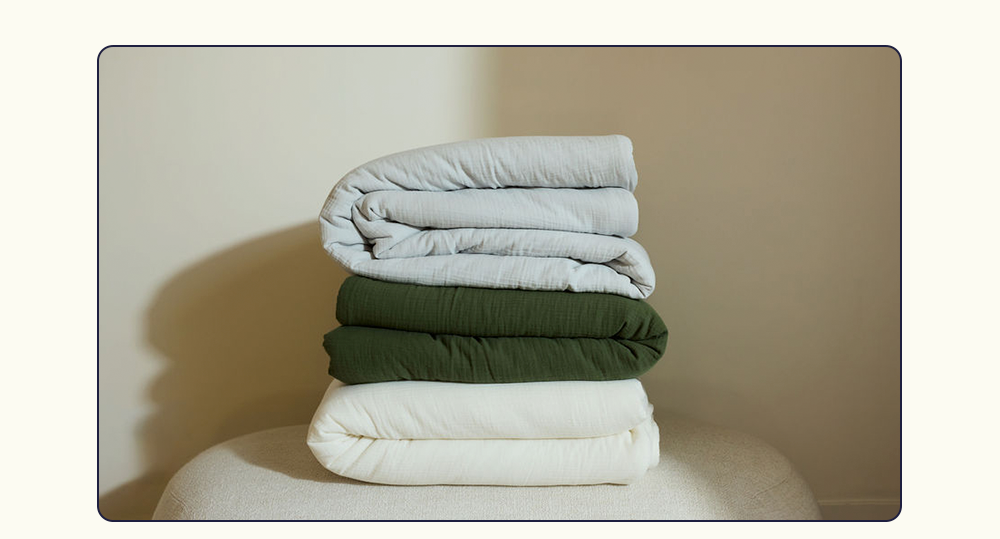 Cotton Quilts in Mist, Moss, and Pearl stacked on each other on a bench.