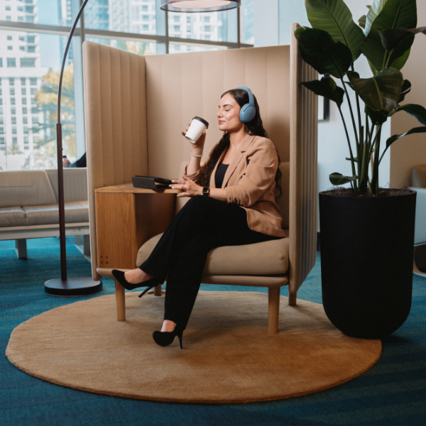 A woman sits in a contemporary office lounge chair, wearing blue over-ear headphones and holding a takeaway coffee cup.