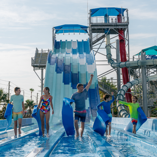 A group of people walk through shallow water at the base of a multi-lane water slide, carrying blue foam mats. The scene takes place at an outdoor water park.