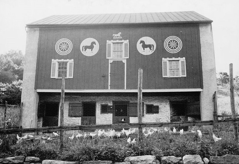 A two-story barn with horse and wheel motifs painted on the upper level, chickens roam in a fenced area in front.
