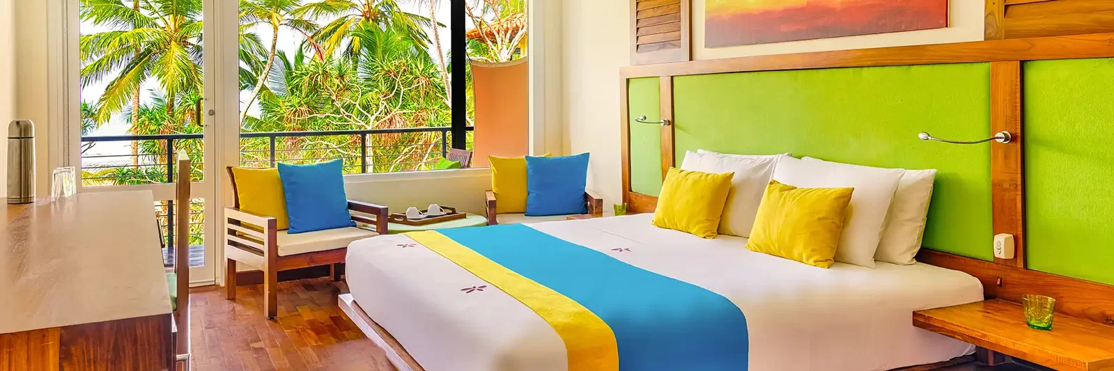 Brightly decorated hotel room with a large bed, colourful cushions, and a view of palm trees outside.