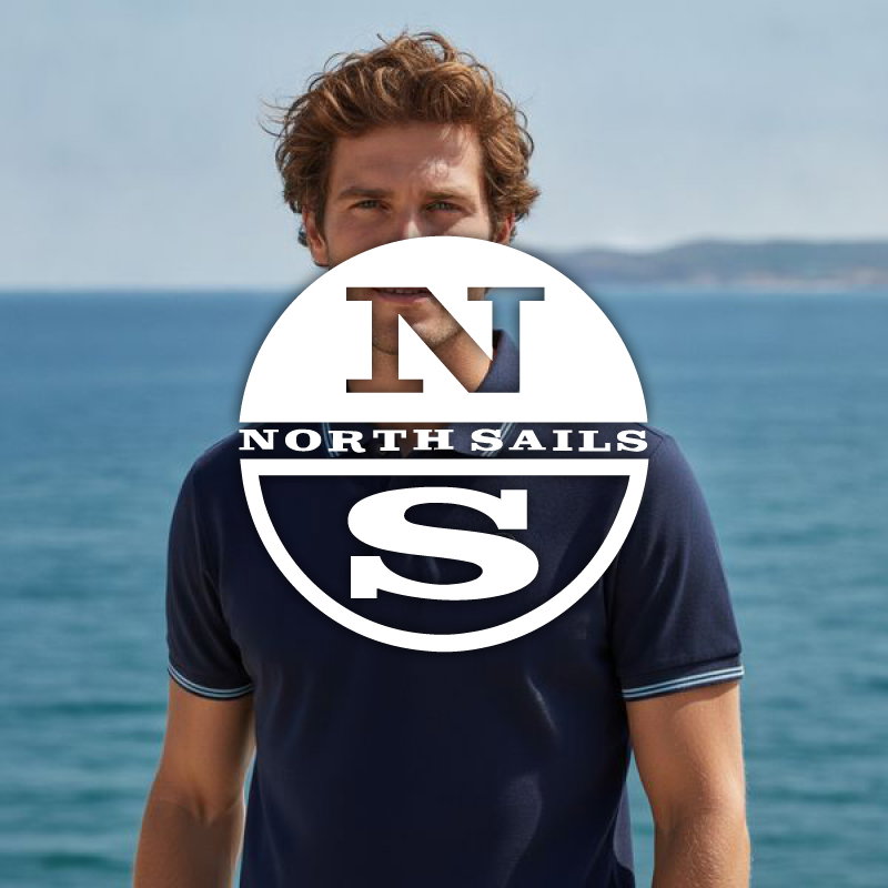 Male model wearing a navy polo shirt with a large North Sails logo overlay, standing by a calm sea coastline