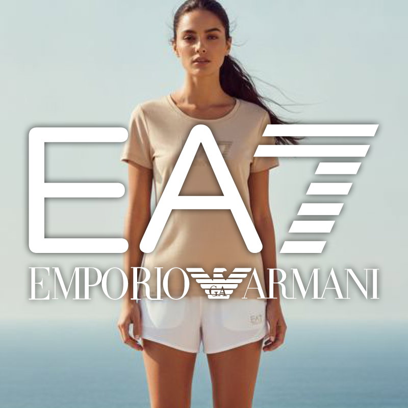 Woman wearing beige T‑shirt and white shorts standing by the sea with a large white EA7 Emporio Armani logo overlaid