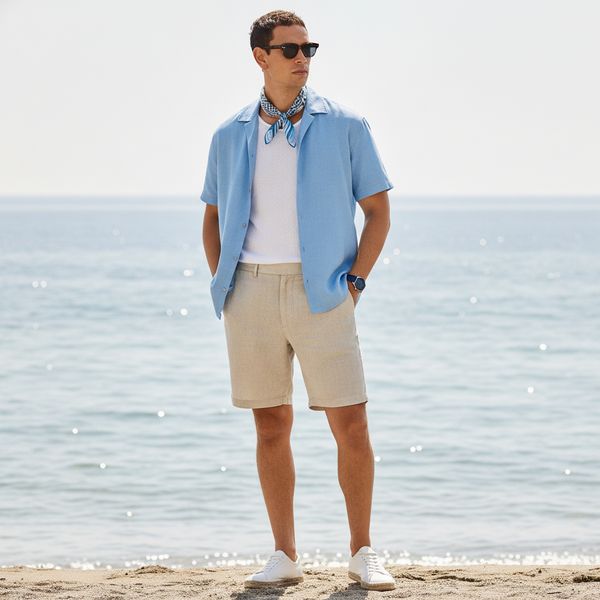Man in light blue open shirt, white T‑shirt, beige shorts and neckerchief standing on sandy beach by the sea