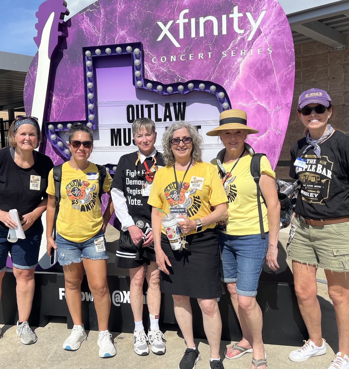 Powered by People volunteers standing in a line smiling at the camera. They're standing in front of a purple guitar pick-shaped Outlaw Music Fest sign 