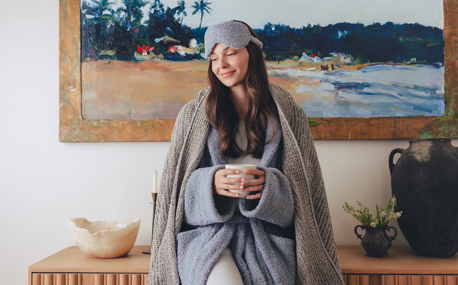 Model wrapped in a Kashwére Lightweight Throw, wearing a soft sleep mask and holding a warm mug indoors. Designed for effortless, breathable comfort, the Lightweight Throw delivers the signature Kashwére softness you love in a lighter layer you'll reach for year-round.