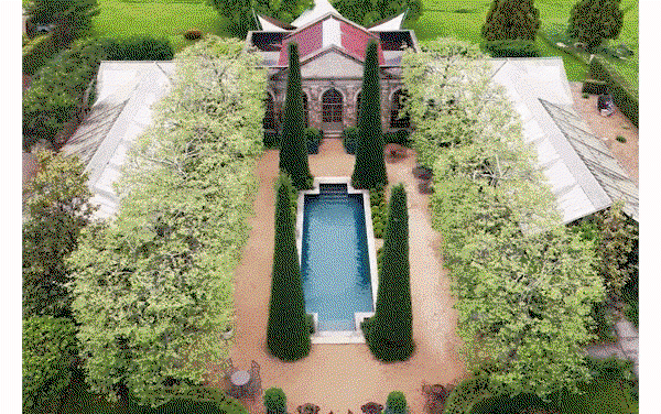 Aerial view of an estate with a rectangular pool, tall cypress trees, and a stone building with a red roof surrounded by lush greenery