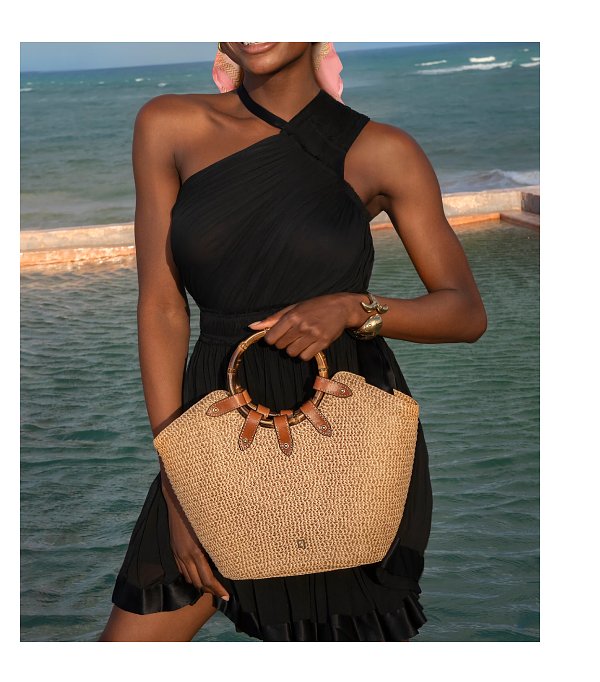 A woman in a black dress holding a woven handbag by the water.