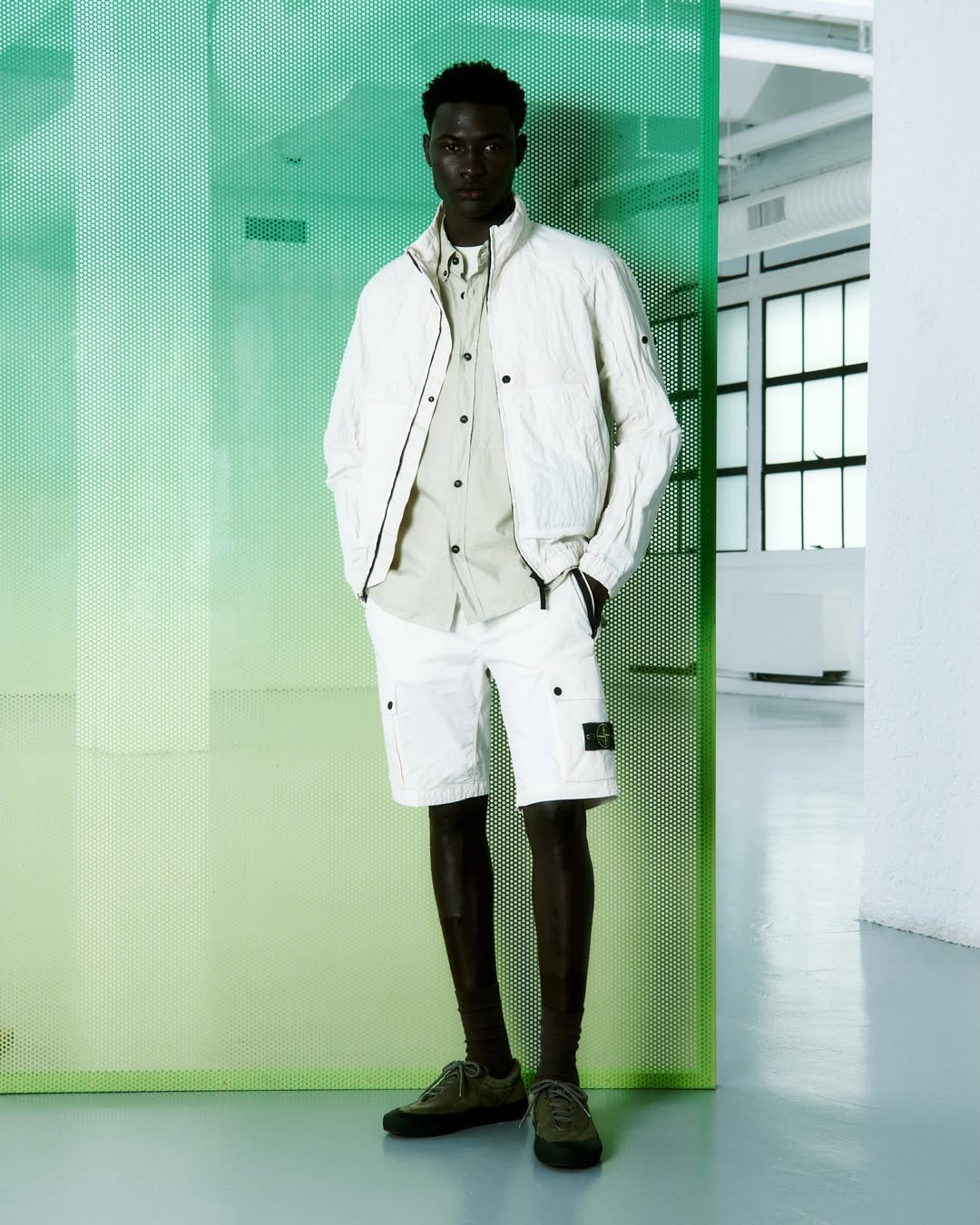 Man wearing white jacket, beige shirt and white cargo shorts with green shoes, standing against a green perforated panel in a bright studio