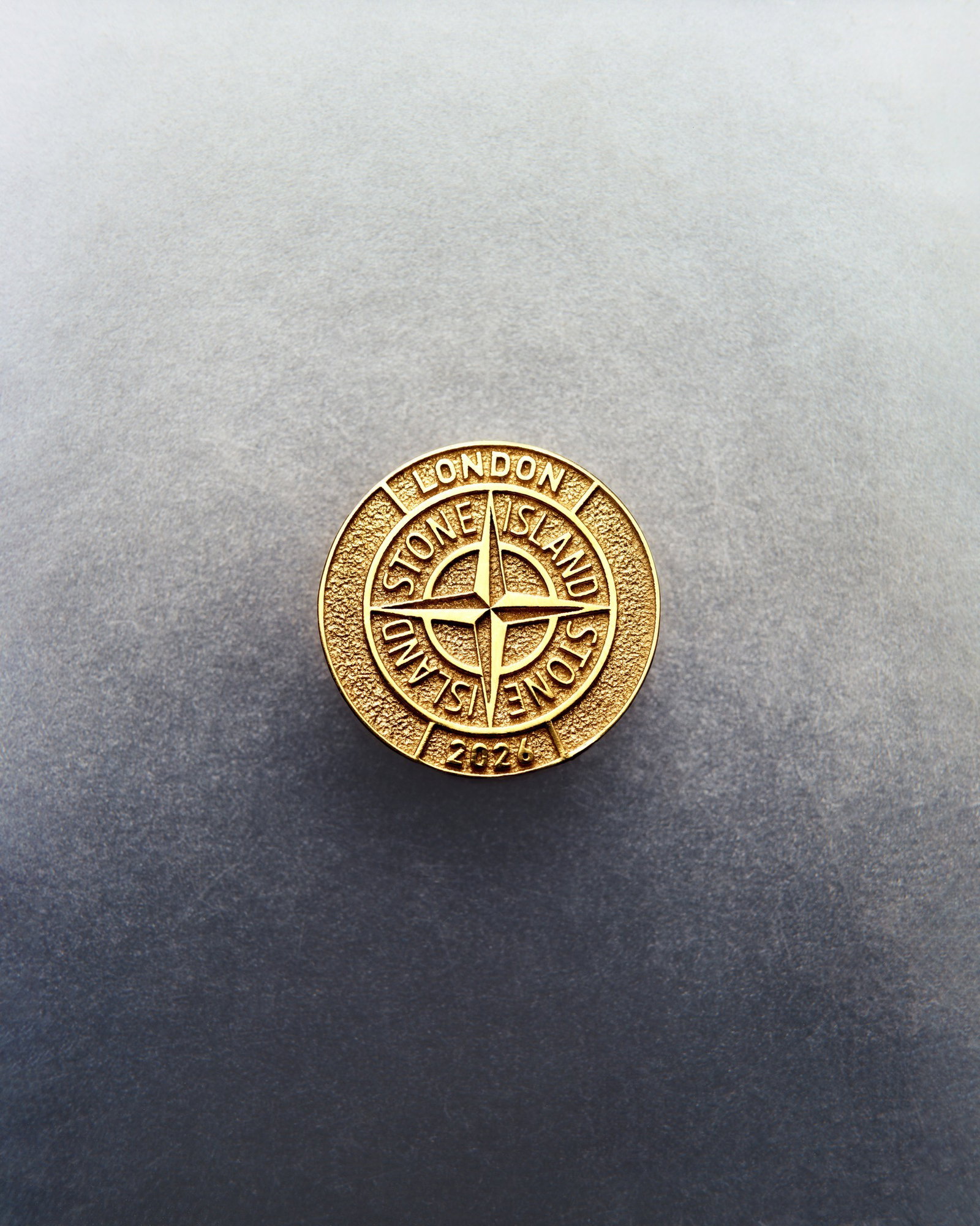 Gold Stone Island round badge with compass logo and 'London' inscription on textured grey background
