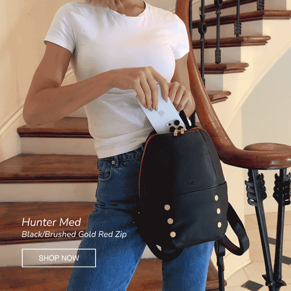 Model placing everyday essentials into the Hammitt Hunter Med backpack in Black/Brushed Gold Red Zip, highlighting its structured silhouette and spacious interior.