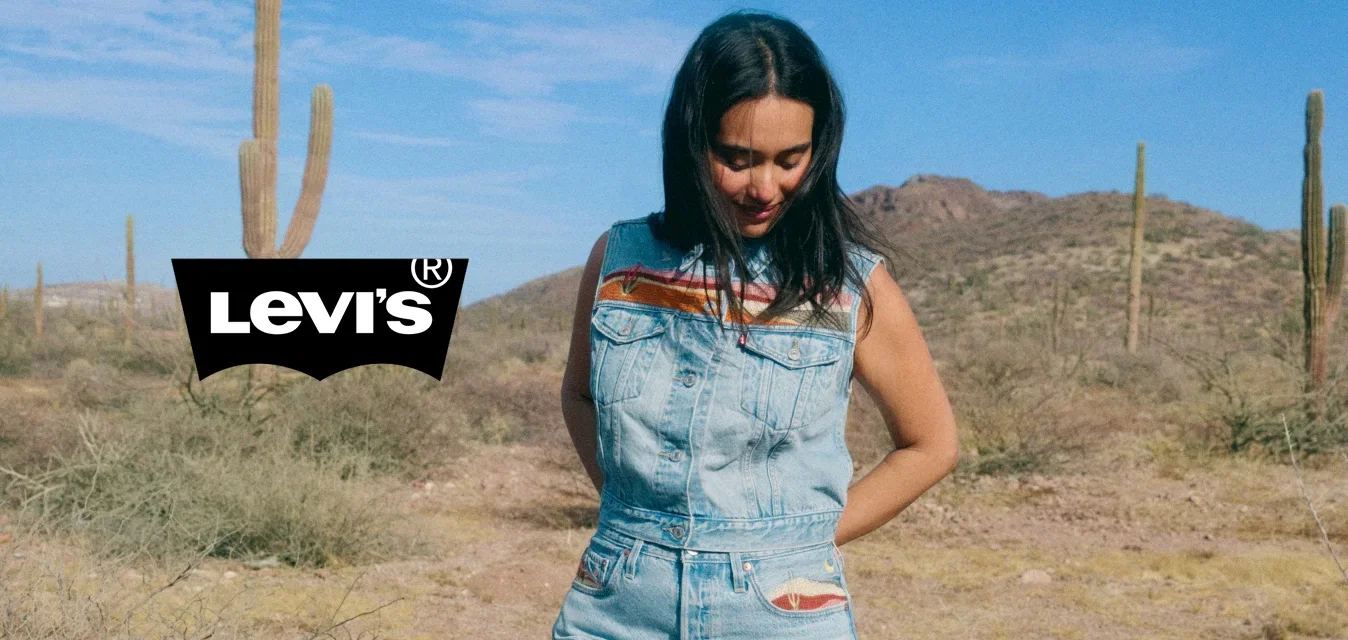 Donna con tuta corta in denim senza maniche in piedi in un deserto con cactus e montagne sotto un cielo azzurro limpido.