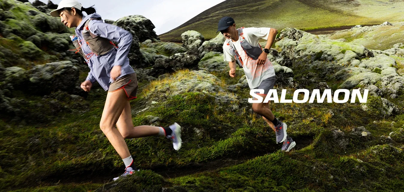 Due corridori trail con abbigliamento sportivo percorrono un pendio roccioso e muschioso con una montagna sullo sfondo.