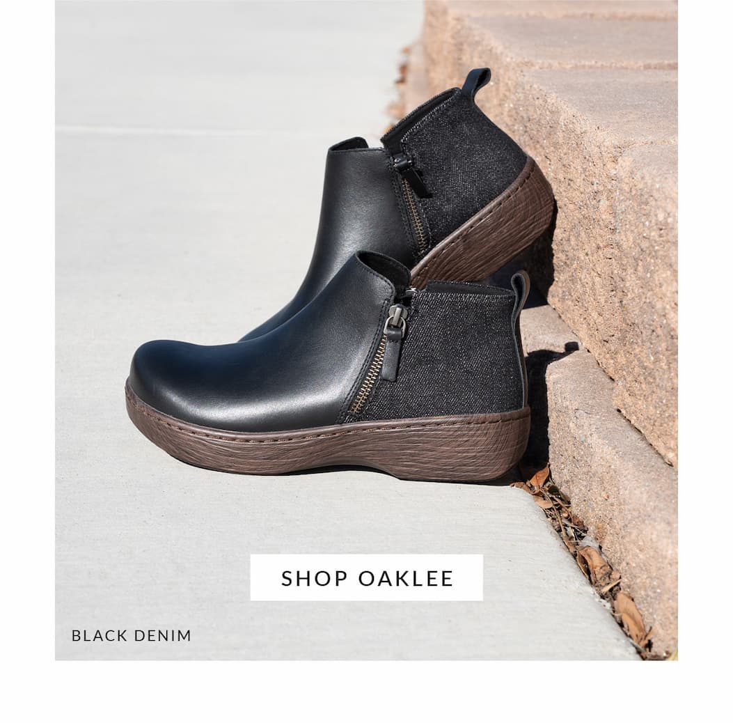A pair of black Alegria Oaklee ankle boots with mixed leather and denim textures and side zippers, resting on concrete sidewalk steps. Features a wood-grain textured sole and a button that reads 'SHOP OAKLEE'.
