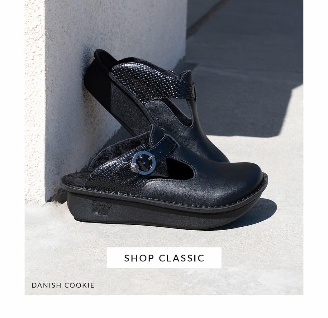 A side view of black Alegria Danish Cookie clog-style shoes leaning against a white textured wall on a concrete surface. The shoes feature a strap with a metallic buckle. A button reads 'SHOP CLASSIC'.