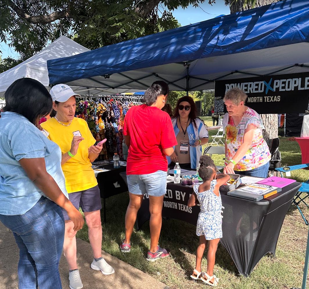 Group of people standing around a Powered by People booth outside. Two people furthest left are looking at a phone, a person in a red shirt is facing the table with a child standing next to them, and there are two people behind the booth table speaking with both of them.
