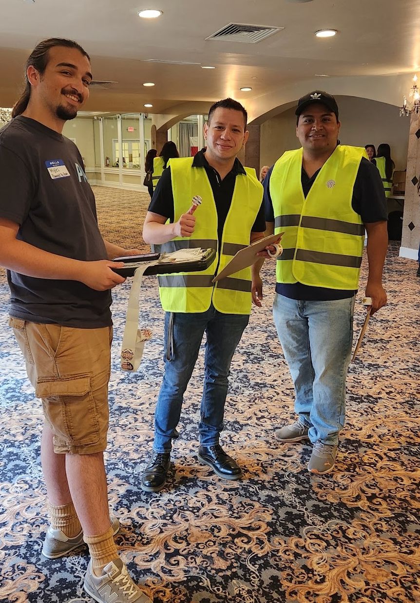 Three Powered by People volunteers, one wearing a PXP tee shirt on left and two wearing high visibility vests on right. They're all holding clipboards and smiling at the camera.