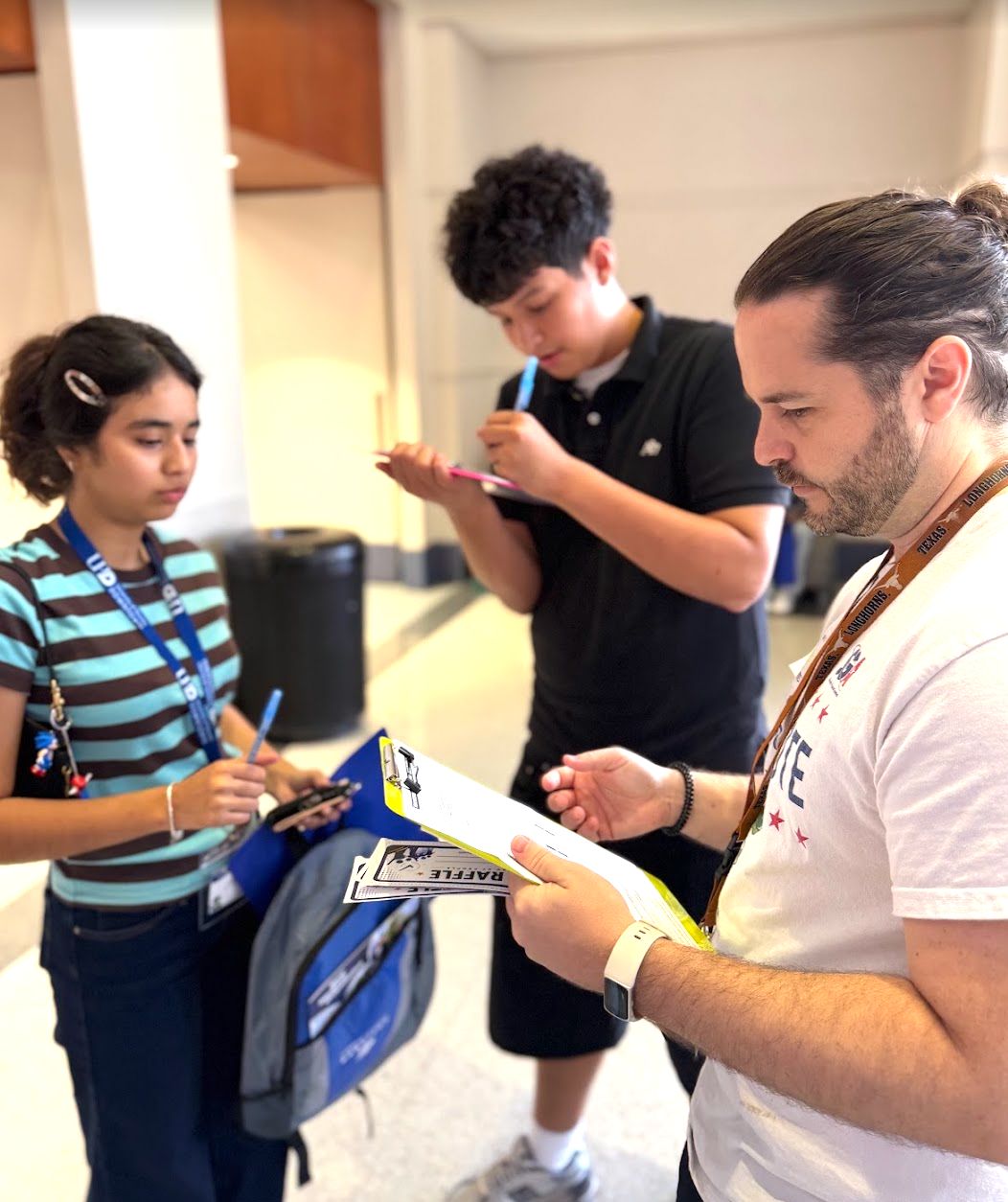 (L-R) Someone wearing a lanyard holding their phone and book bag, student filling out their voter registration form on a clipboard, someone holding a clipboard and raffle tickets facing towards the other two people