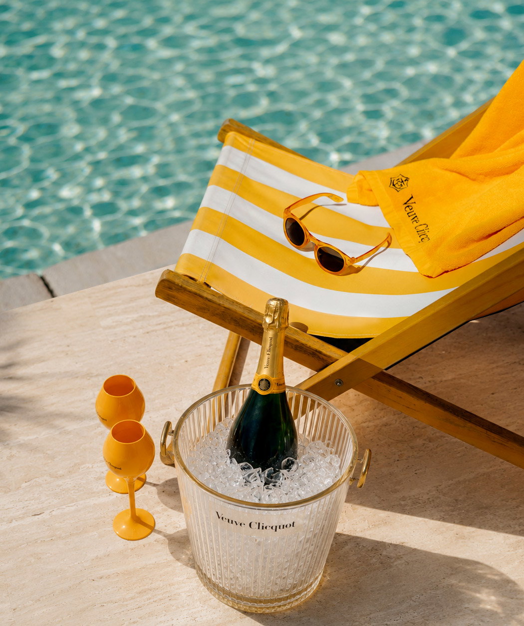 Poolside scene with Veuve Clicquot champagne chilling in an ice bucket beside a yellow-striped deckchair, towel, sunglasses and two yellow flutes