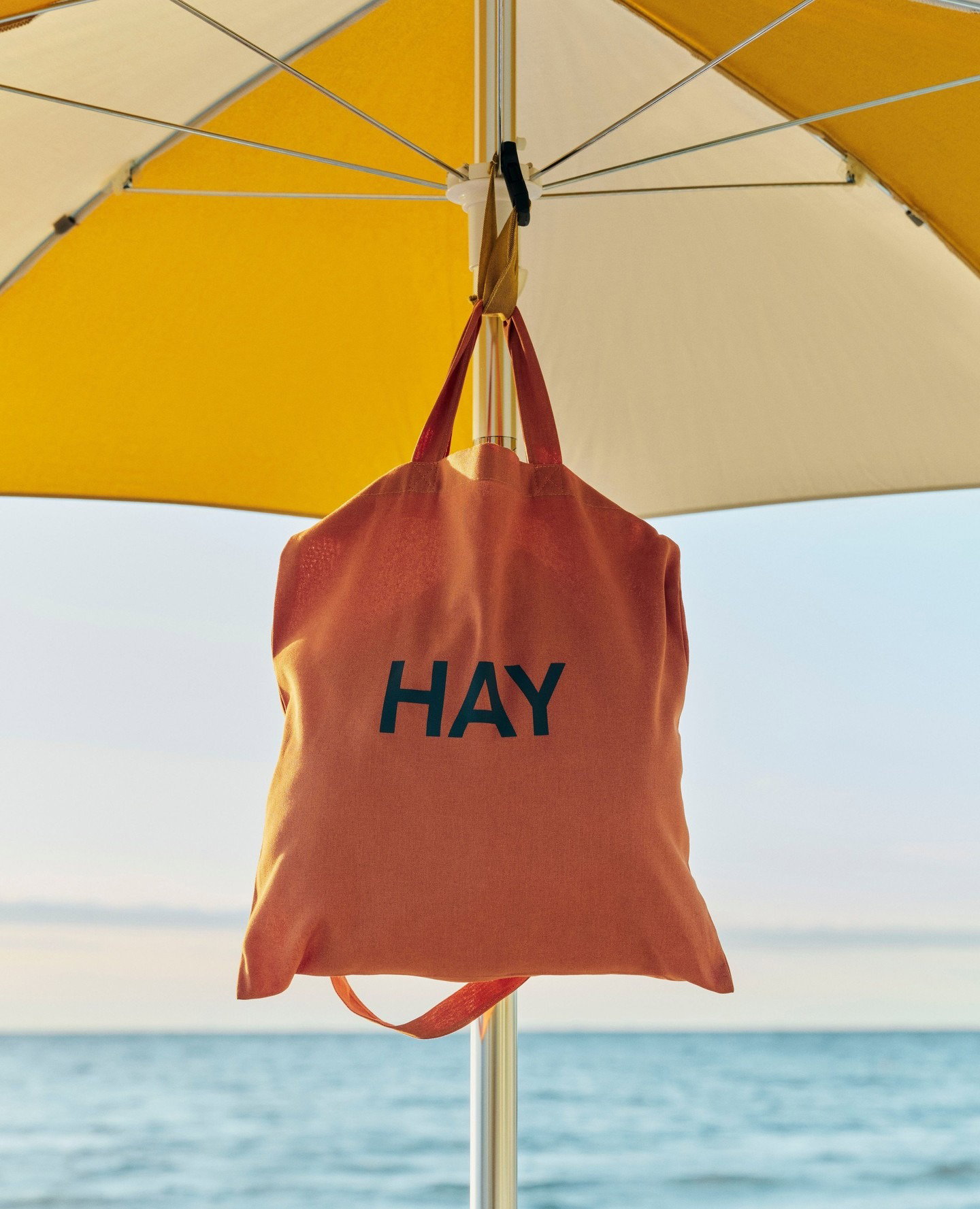 Orange HAY-branded canvas tote bag hanging from a yellow beach umbrella pole with calm sea and sky in the background
