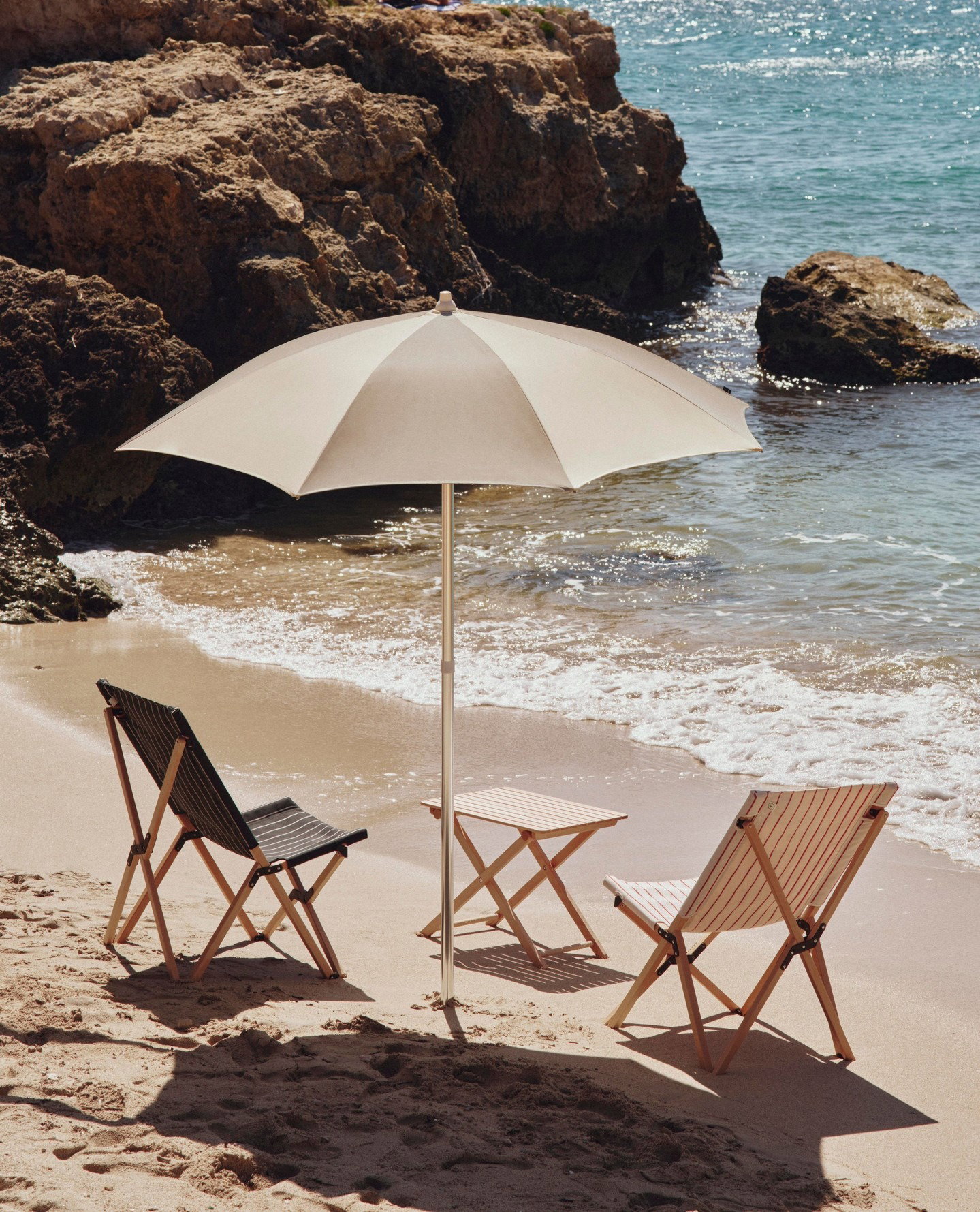 Three wooden folding beach chairs and a white parasol on a sunlit sandy shore beside rocky coastline and turquoise sea