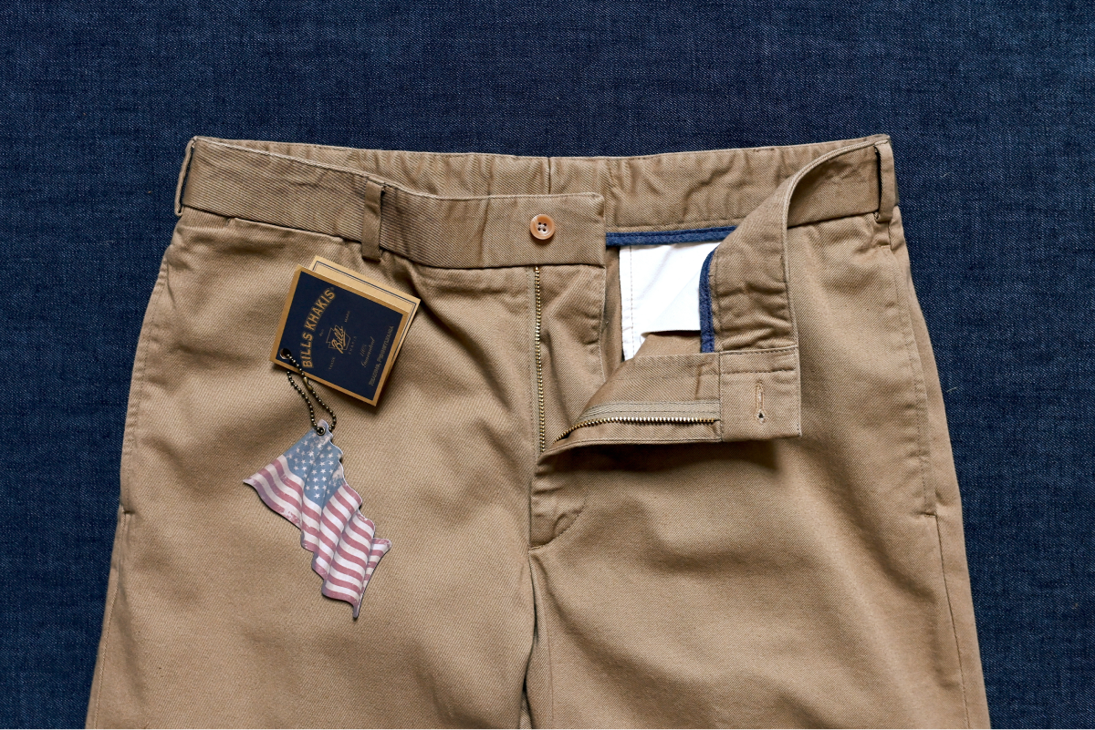 A pair of Bills Khakis khaki pants lay on a blue surface with waiste slightly open. The iconic Bills Khakis tags are on display