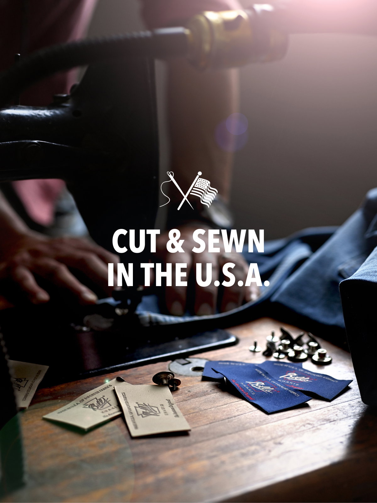 Hands work on a vintage sewing maching dewing Bills Khakis fabric tags with a dramatic lighting. Text reads Cut and Sewn in the USA with a needle and flag icon