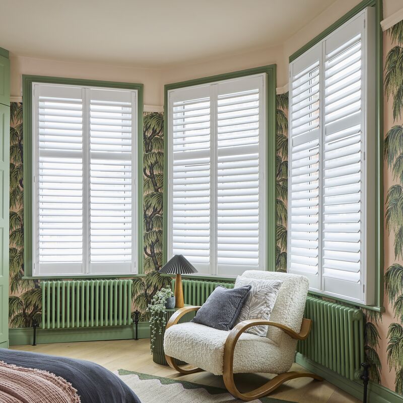 Bay window with white shutters framed by green trim, softly filtering light in a bedroom with botanical wallpaper and cosy chair.