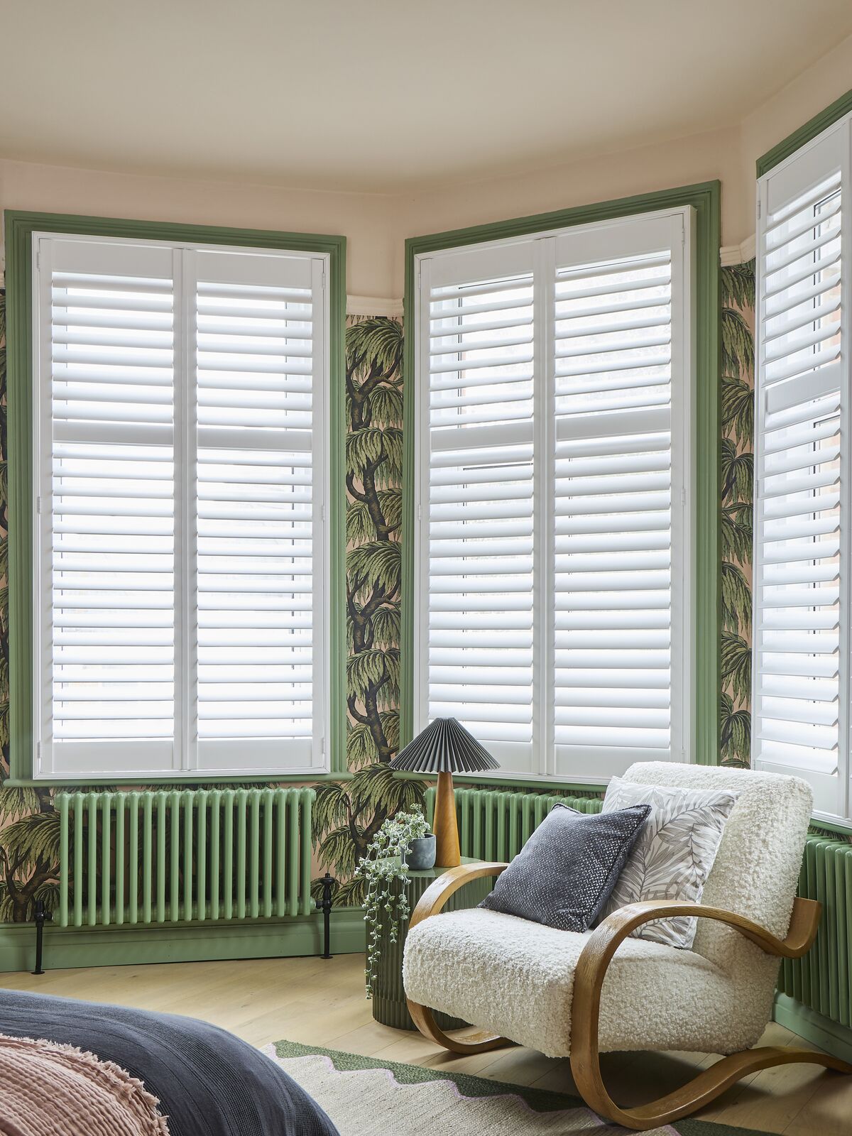 Bay window with white shutters framed by green trim, softly filtering light in a bedroom with botanical wallpaper and cosy chair.