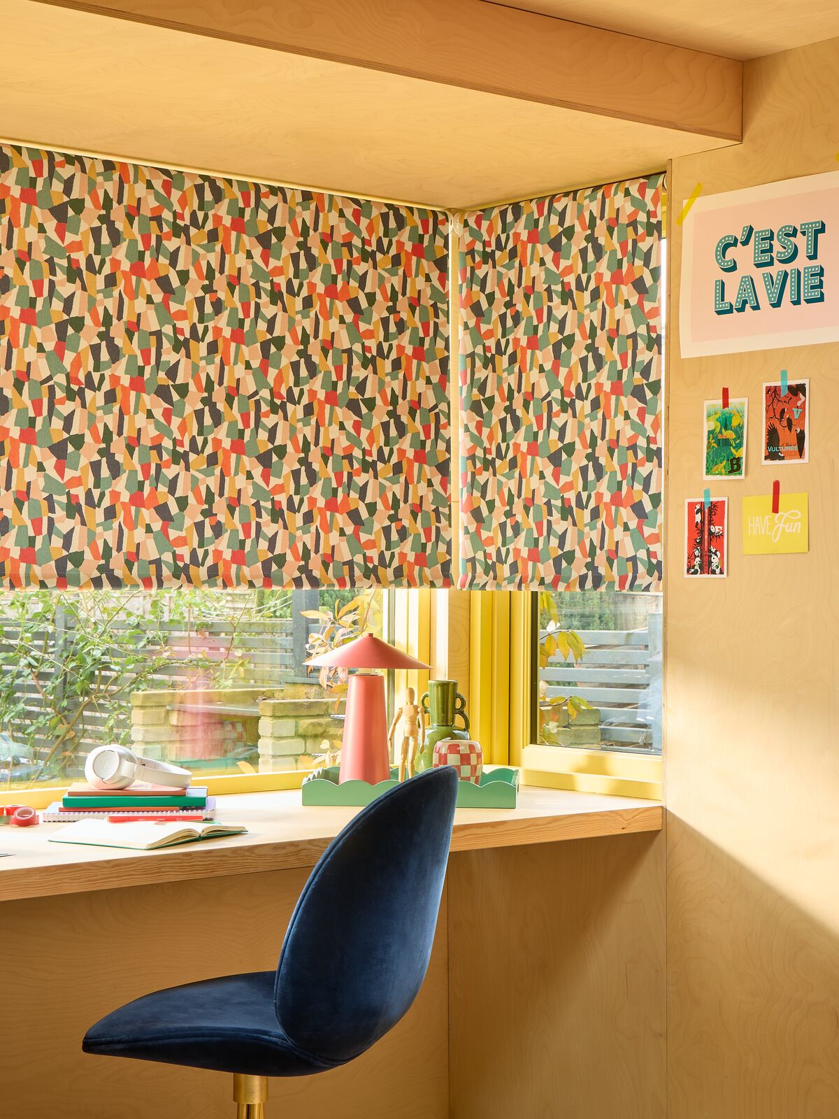 Corner window with vibrant patterned Roller blinds, casting warm light over a desk setup with stationery and garden views beyond.