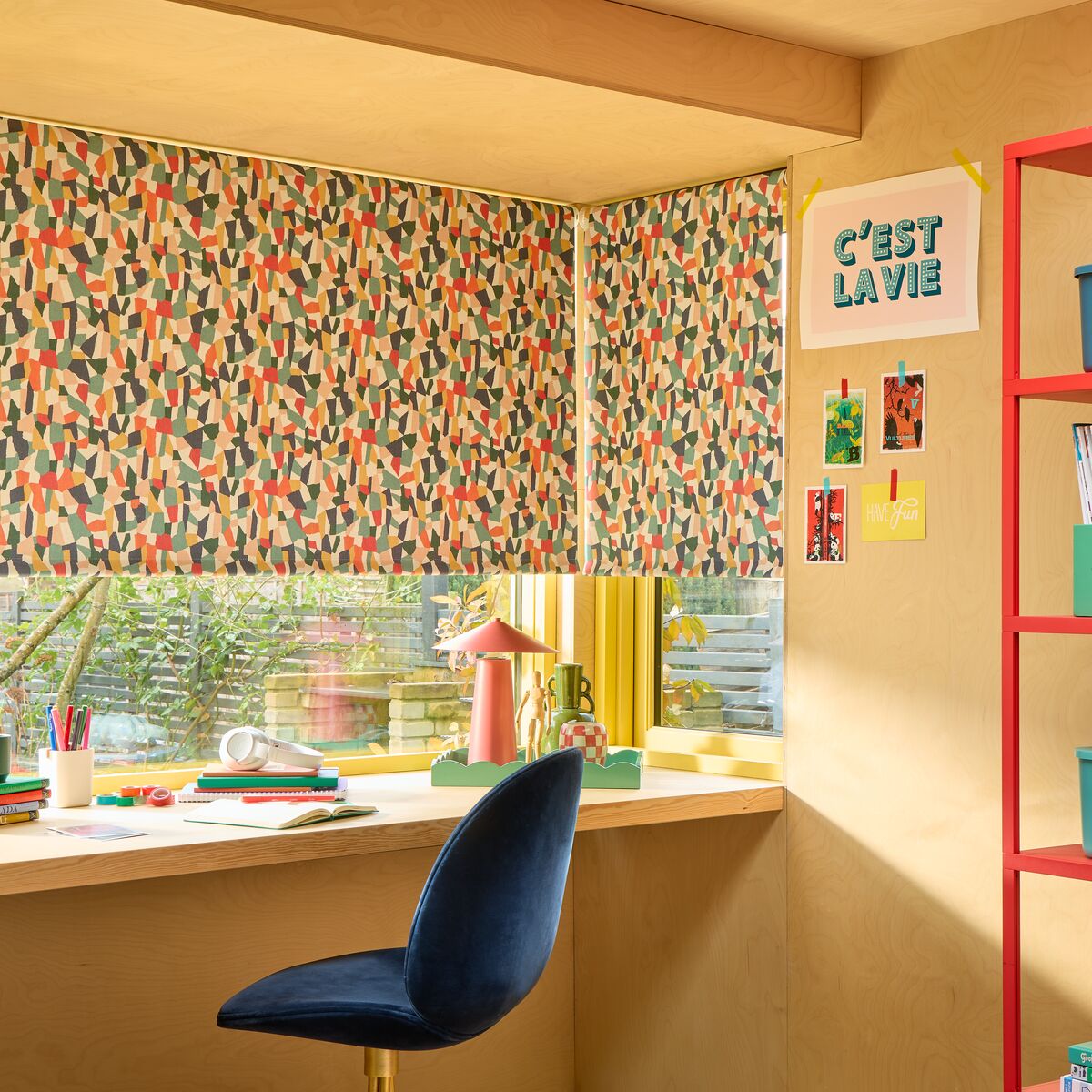 Corner window with vibrant patterned Roller blinds, casting warm light over a desk setup with stationery and garden views beyond.