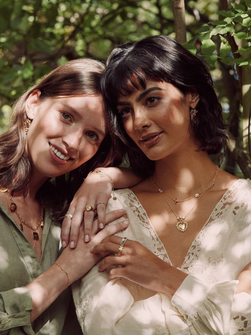 Two models wearing layered gold necklaces and rings from the Summer Favourites collection, against a summer lit forest background.