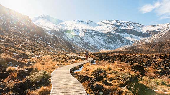 Frau läuft über Holzsteg durch Tongariro Nationalpark mit schneebedeckten Gipfelbergen.