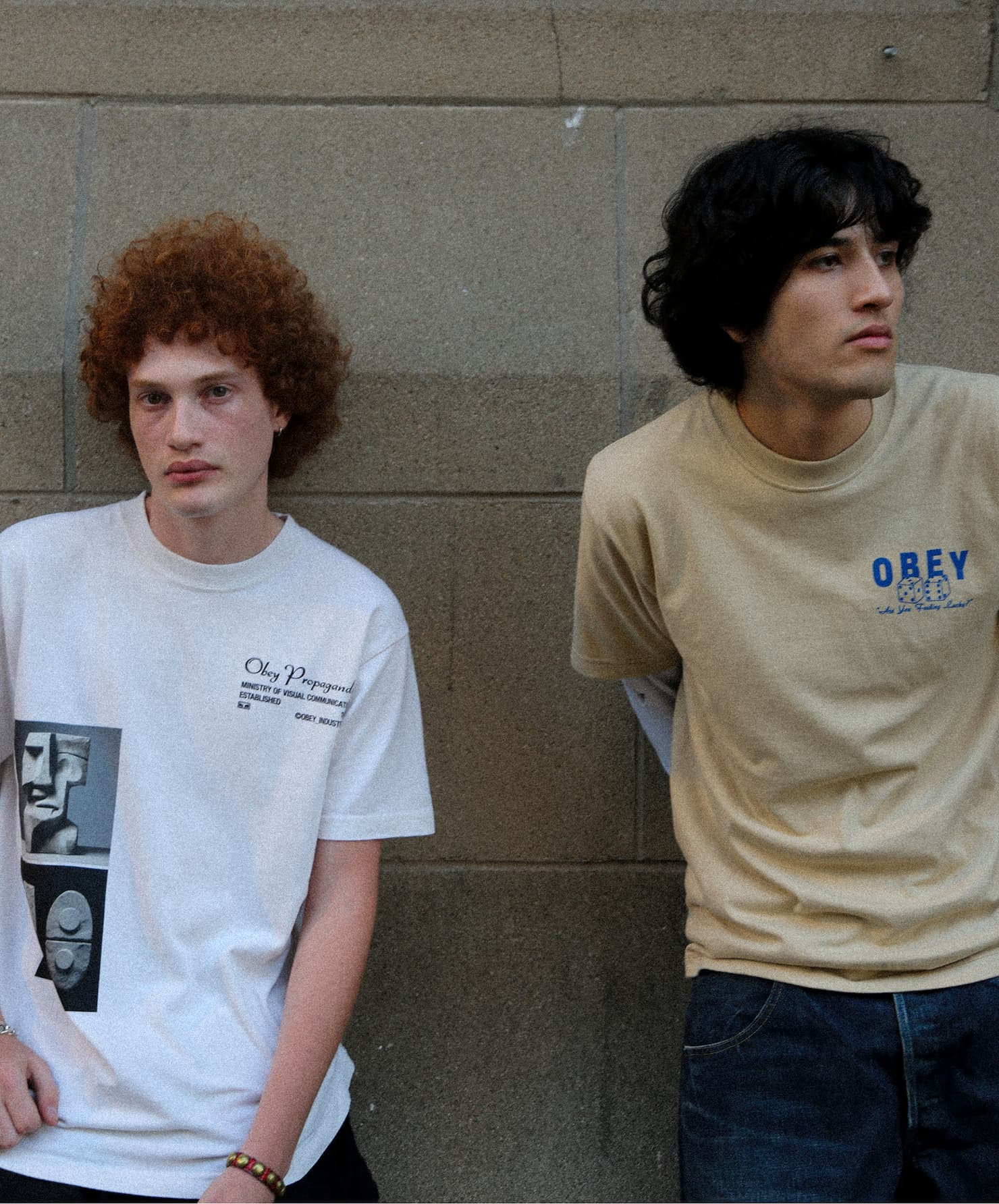 Two young men posing against a stone wall showcasing Obey graphic t-shirts.