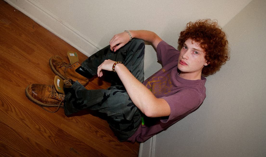 Model sitting against a beige wall wearing a muted purple Obey t-shirt, dark cargo pants, and work boots.