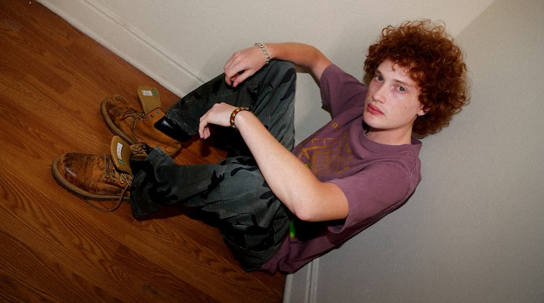 Model sitting against a beige wall wearing a muted purple Obey t-shirt, dark cargo pants, and work boots.