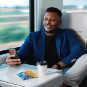 A man sits comfortably by the window on a modern train, looking at his smartphone and smiling. He wears a blue blazer and light pants, seated at a white table with a coffee cup and a small snack packet. The blurred landscape outside the window suggests the train is moving at speed. The interior appears clean and contemporary, creating a relaxed, productive travel atmosphere.