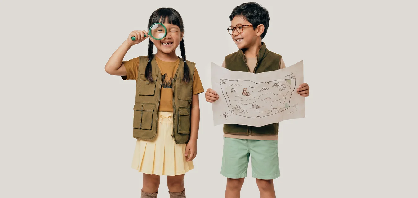 Deux enfants portant des tenues d'explorateurs, l'un tenant une loupe et l'autre une carte au trésor, tous deux souriants.