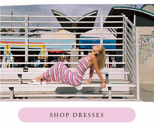 Model on bleachers wearing pink striped dress. Shop dresses. 