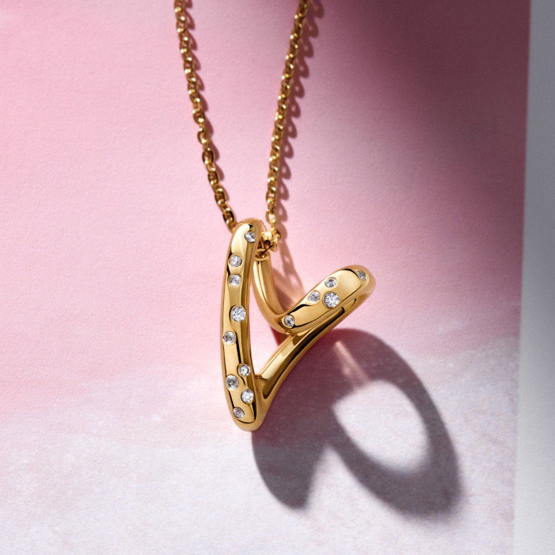 A close-up of a 14k gold-plated heart-shaped necklace on a pink background.