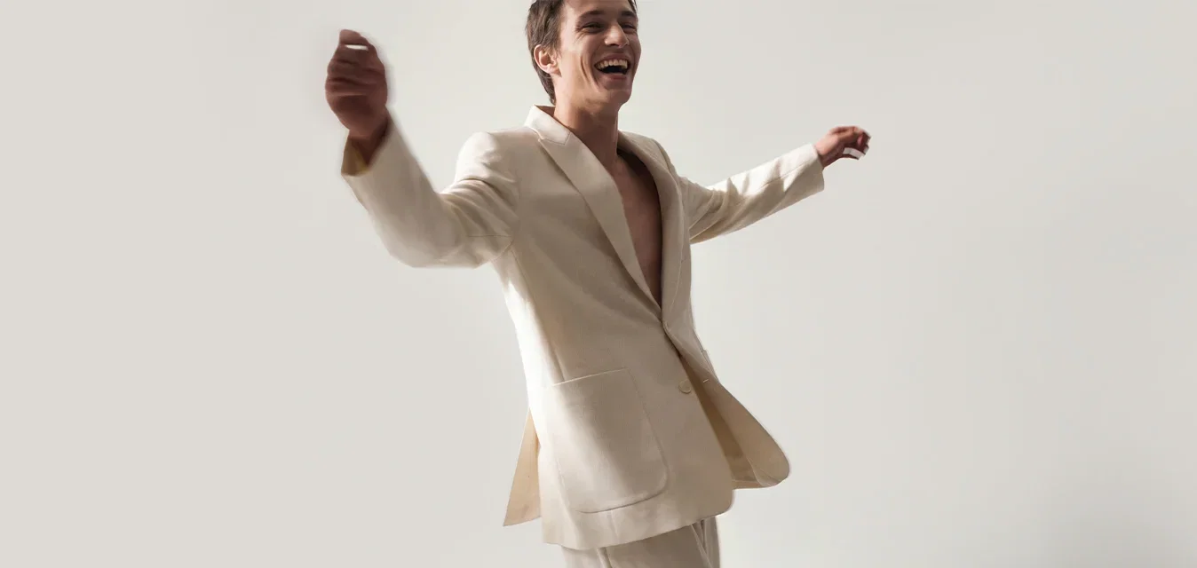 Hombre con un traje beige claro sonríe y levanta los brazos mientras camina frente a un fondo gris claro liso.