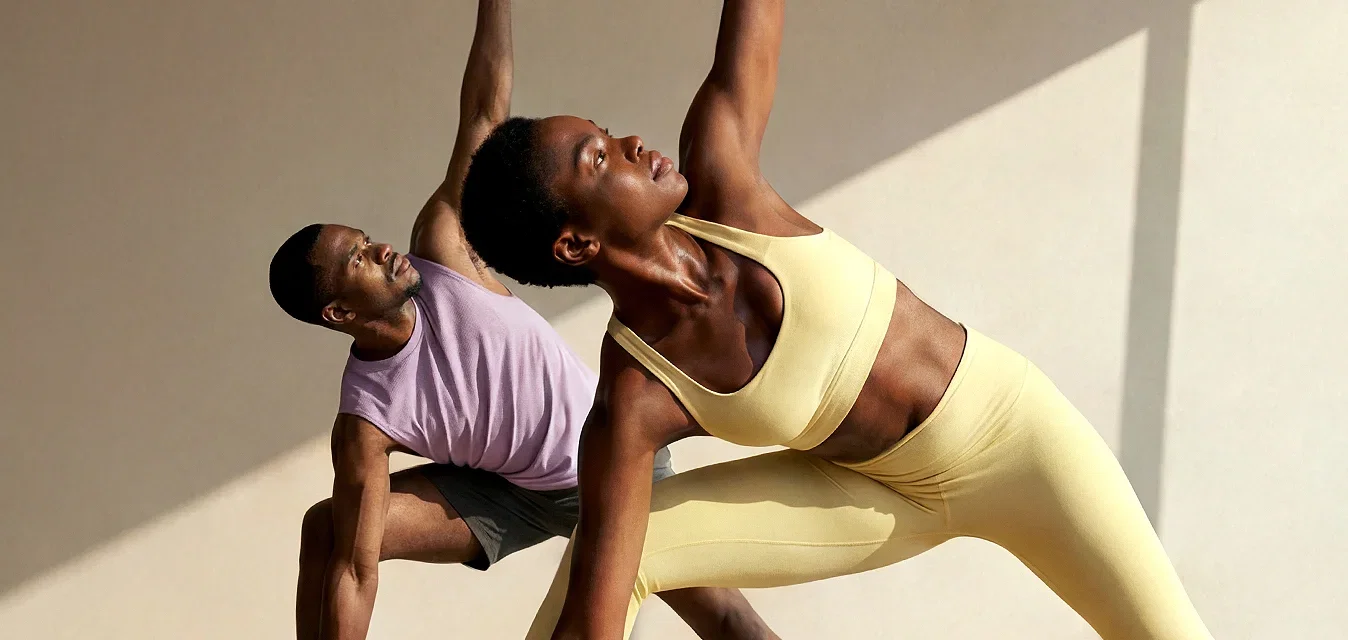 Dos personas practicando yoga sobre esterillas en una habitación soleada con grandes ventanas, ambas en la postura de ángulo lateral extendido.